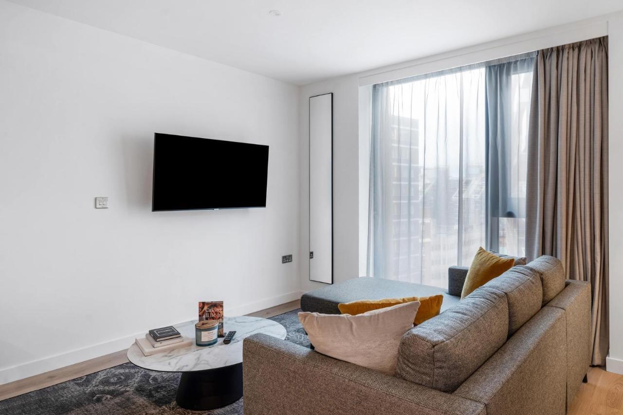 Citysuites 2 Aparthotel Manchester Exterior foto The image shows a modern living room with a minimalist design. There is a large flat-screen TV mounted on the white wall. A comfortable gray sofa with textured cushions occupies the center of the room, complemented by a round coffee table in front of