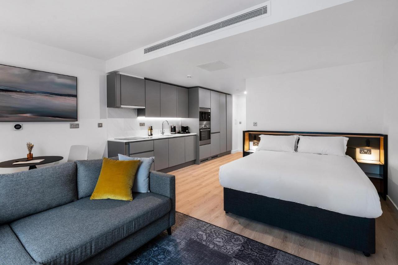 Citysuites 2 Aparthotel Manchester Exterior foto The photo shows a modern studio apartment. On one side, there is a comfortable gray sofa adorned with a yellow cushion. Nearby, a round table is placed, likely intended for dining or casual sitting. 

The kitchen area features sleek gray cabinetry wi