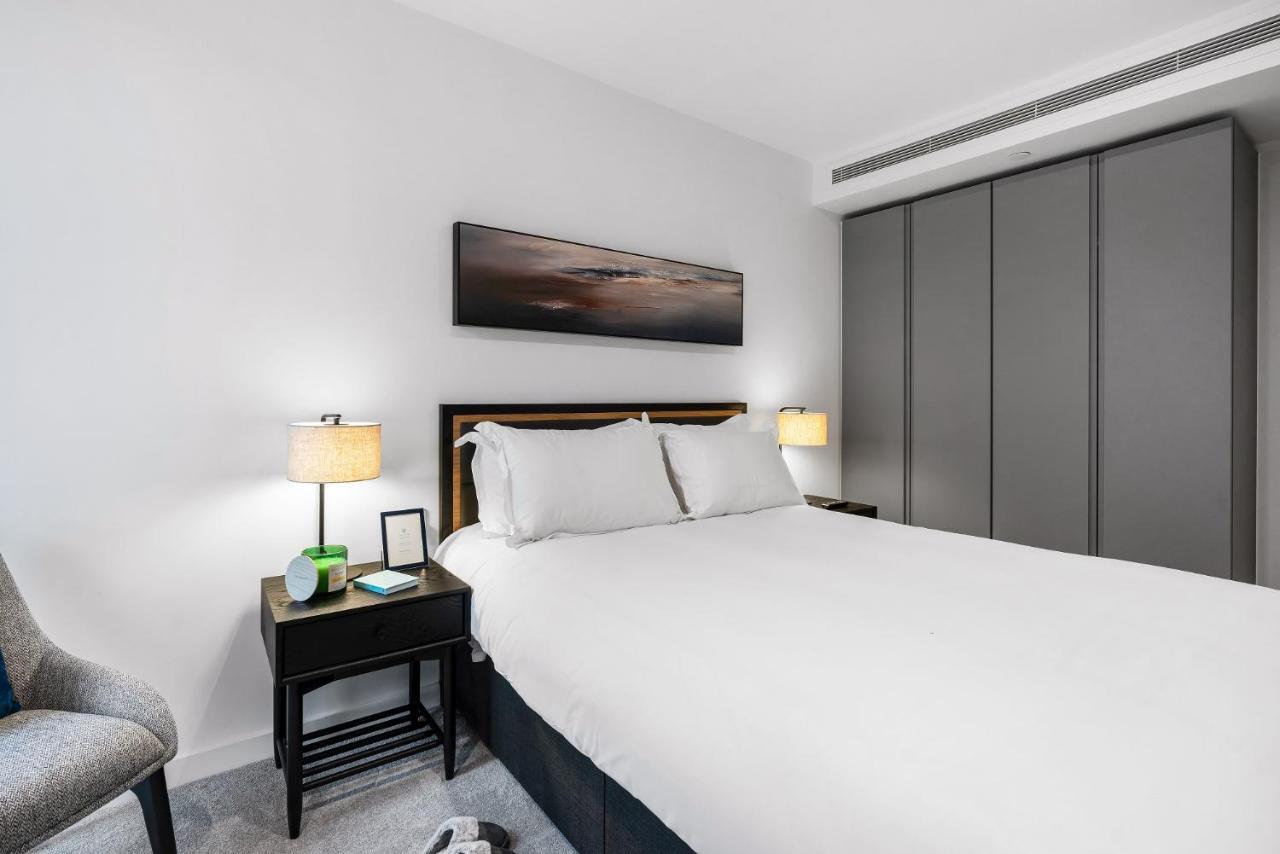Citysuites 2 Aparthotel Manchester Exterior foto The photo shows a modern bedroom with a neatly made bed featuring white linens and pillows. Flanking the bed are two bedside tables, each with a lamp providing soft lighting. There is a piece of artwork above the bed, which appears to depict a landsc