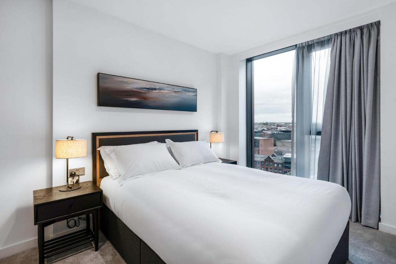 Citysuites 2 Aparthotel Manchester Exterior foto The image shows a modern bedroom featuring a large bed with a white duvet and two pillows. On either side of the bed are small nightstands with lamps. A large window occupies one wall, draped with light gray curtains, allowing natural light to enter 