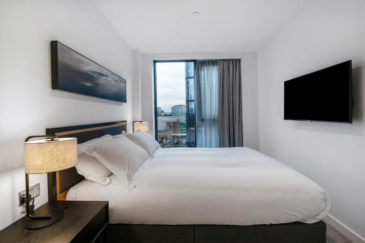 Citysuites 2 Aparthotel Manchester Exterior foto The photo shows a modern bedroom with a minimalist aesthetic. It features a large bed with white bedding and two pillows. On either side of the bed, there are bedside tables with lamps. A large window lets in natural light, covered by sheer curtains.