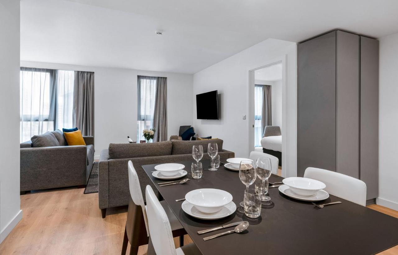 Citysuites 2 Aparthotel Manchester Exterior foto The photo shows a modern living space with an open layout. In the foreground, there is a dining table set for a meal, featuring multiple plates, glasses, and cutlery arranged neatly. In the background, there is a comfortable-looking gray sofa with a 