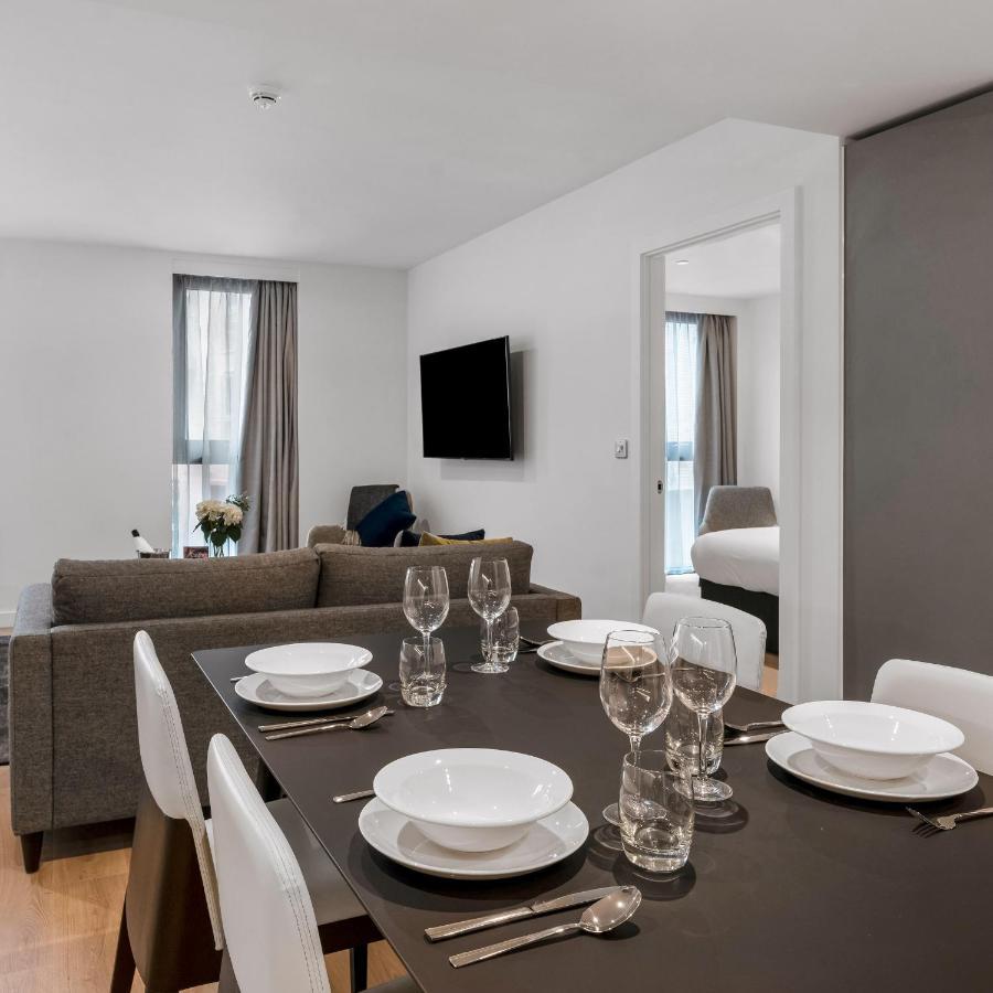 Citysuites 2 Aparthotel Manchester Exterior foto The photo shows a modern, well-lit living space that features a dining area and a sitting area. In the foreground, there is a dining table set for six, with white plates and crystal glasses arranged neatly. The table is surrounded by white chairs. 

