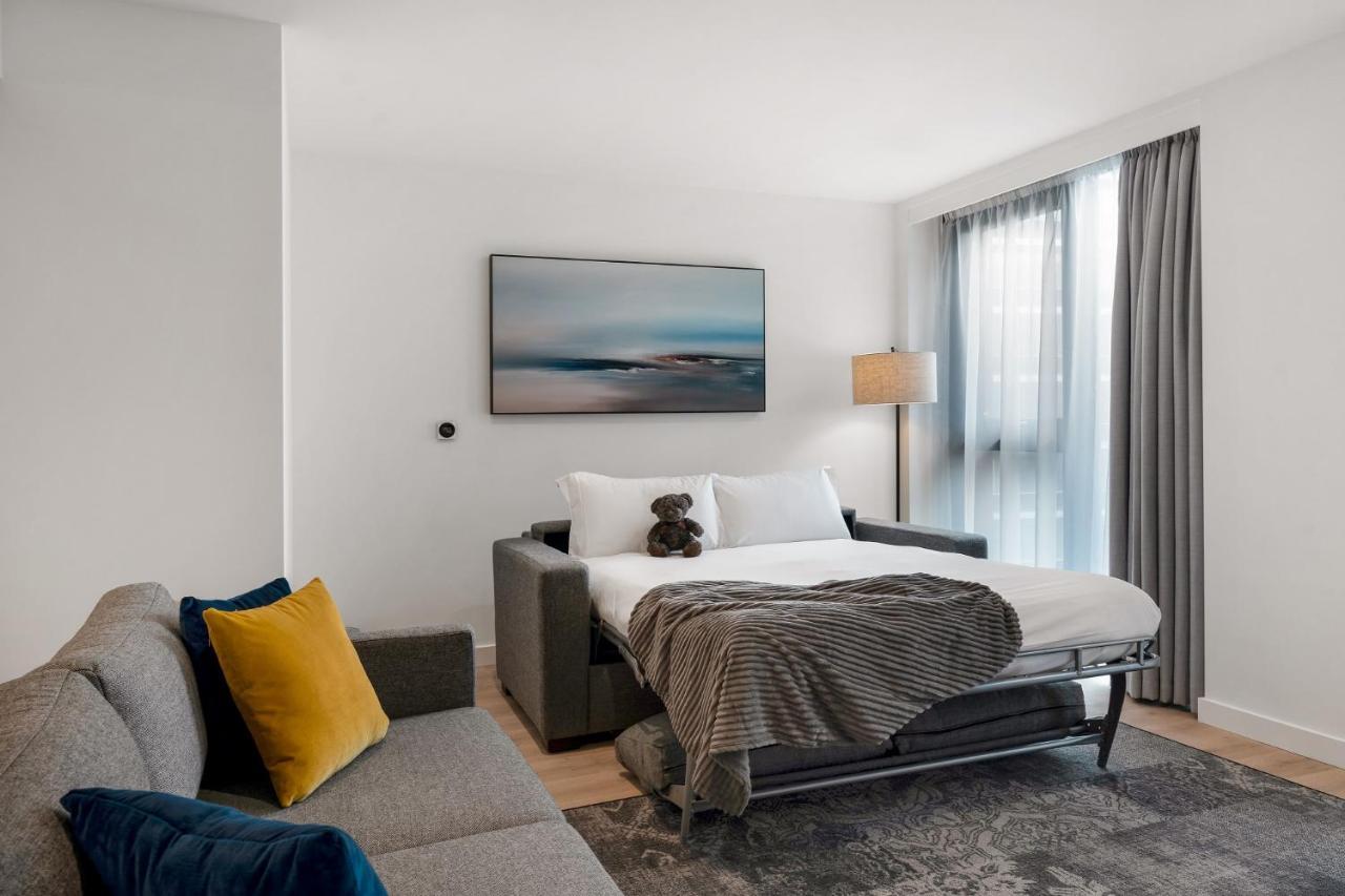 Citysuites 2 Aparthotel Manchester Exterior foto The photo shows a modern and minimalistic guest room. There is a sofa on the left side, adorned with a colorful cushion. In the middle is a bed that appears to be a pull-out style, featuring white bedding and a gray throw blanket. A stuffed toy is pl