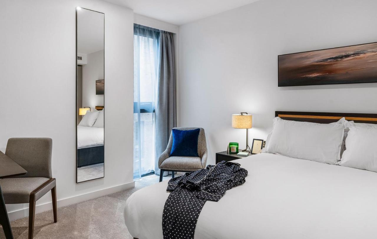 Citysuites 2 Aparthotel Manchester Exterior foto The photo shows a modern hotel room. It features a neatly made bed with white linens and a dark-colored throw draped over it. There is a small bedside table with a lamp next to the bed. A comfortable chair in a light color is positioned near the wind