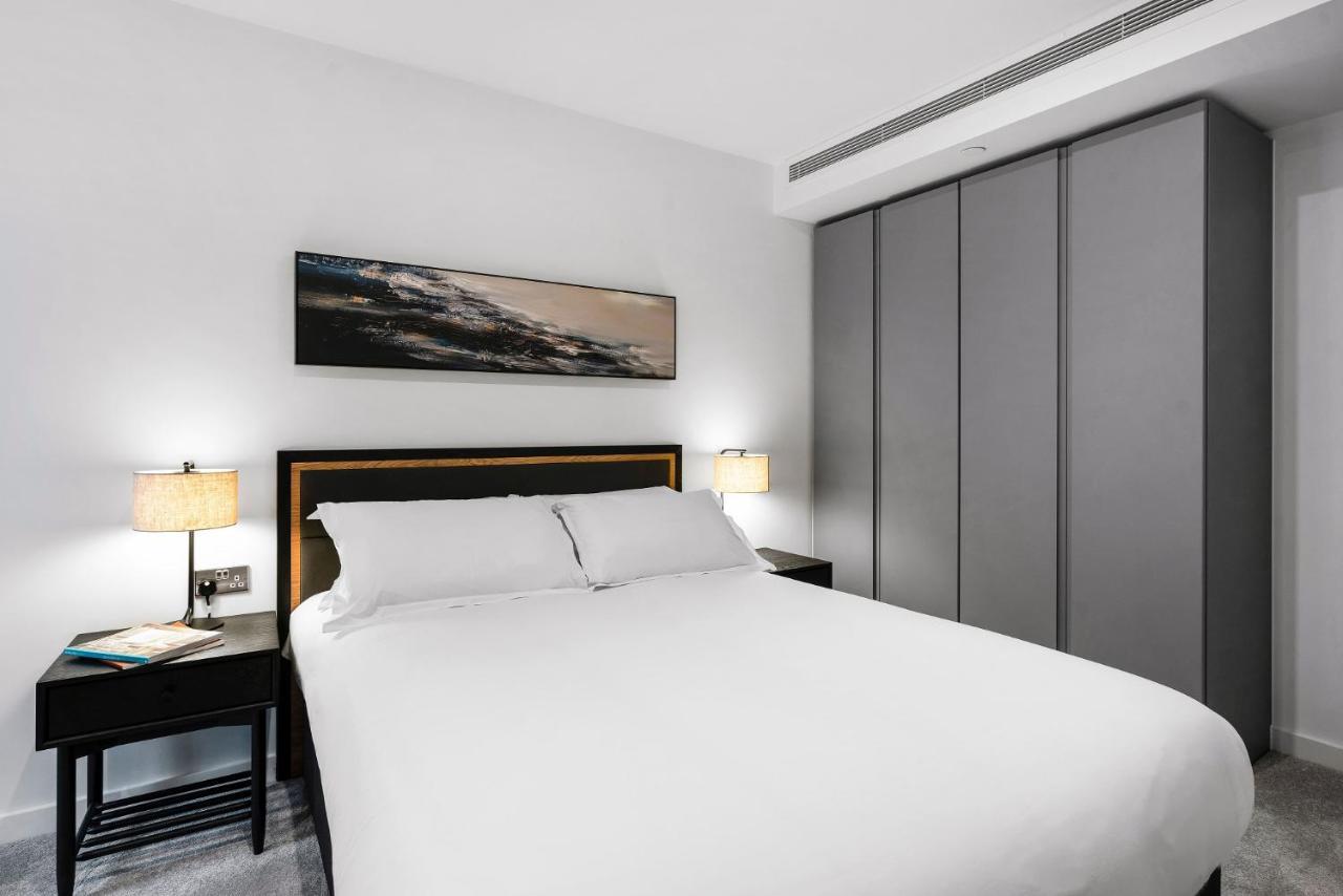 Citysuites 2 Aparthotel Manchester Exterior foto The photo shows a modern bedroom designed with a minimalist aesthetic. The focal point is a large bed with white bedding, positioned against a wall. On either side of the bed, there are black nightstands topped with lamps, providing a warm glow. Abov