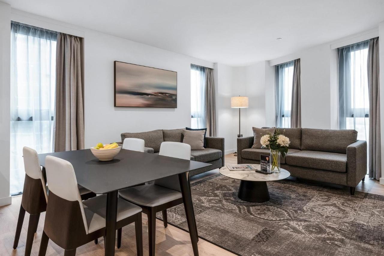 Citysuites 2 Aparthotel Manchester Exterior foto The photo depicts a modern and stylish interior of a living space. On the left, there is a dining table with four chairs, featuring a bowl of fruits on the table. In the background, there's a cozy seating area with a gray sofa and a smaller round cof