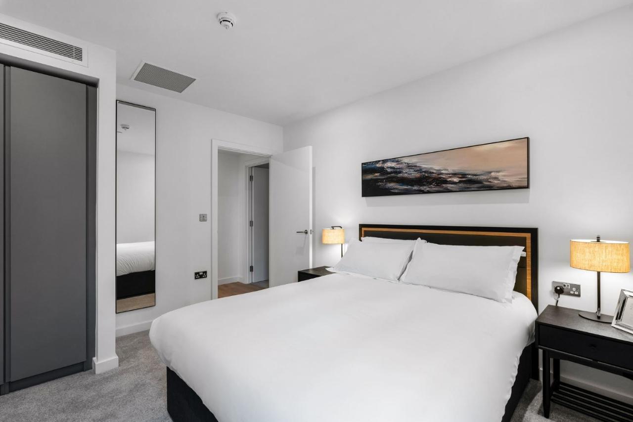 Citysuites 2 Aparthotel Manchester Exterior foto The photo shows a modern bedroom featuring a large double bed with white bedding. There are two bedside tables with lamps on either side of the bed. The walls are painted white, and there's a piece of artwork above the bed that depicts a landscape. A