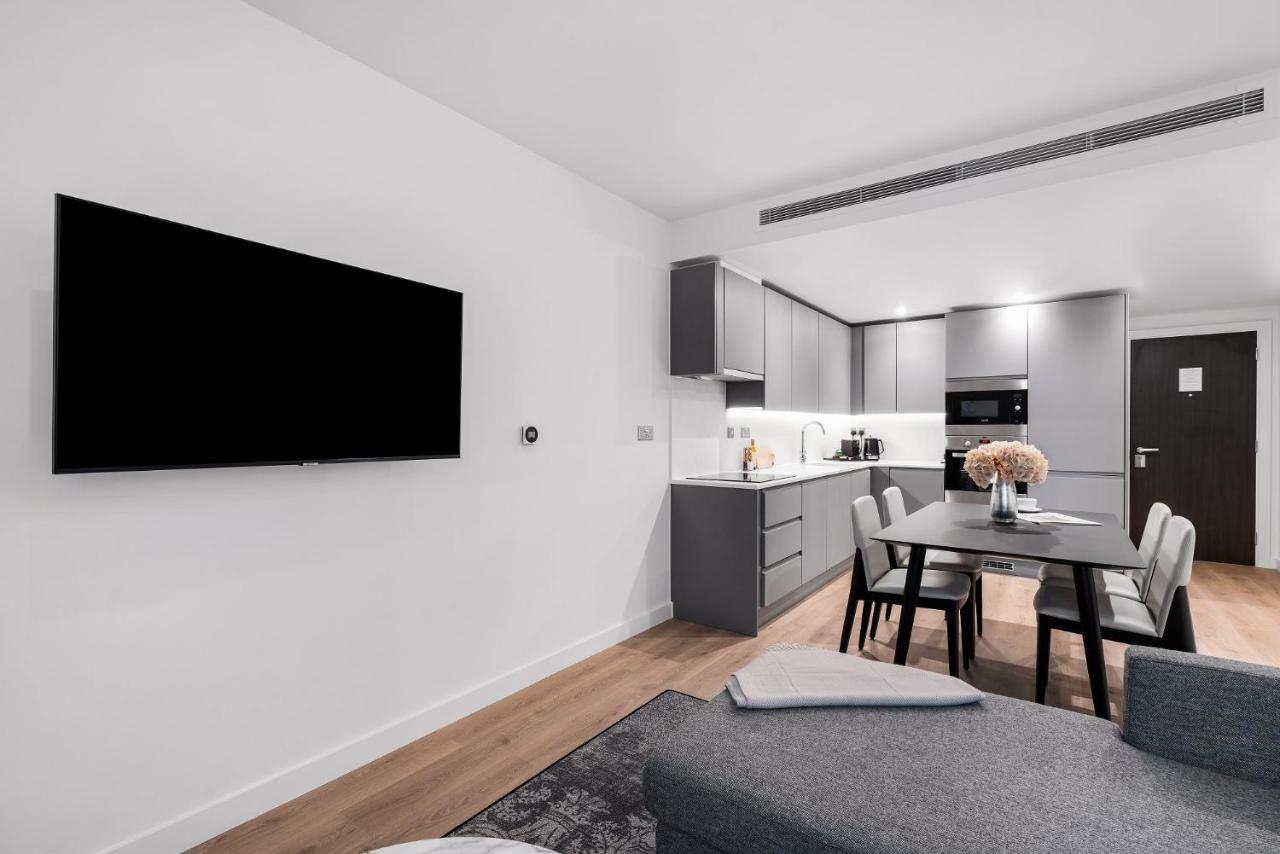Citysuites 2 Aparthotel Manchester Exterior foto The photo shows a modern, minimalist living space. On the left, there is a large flat-screen TV mounted on the wall. To the right, a kitchen area features sleek gray cabinets and appliances, including a built-in microwave. In front of the kitchen, th