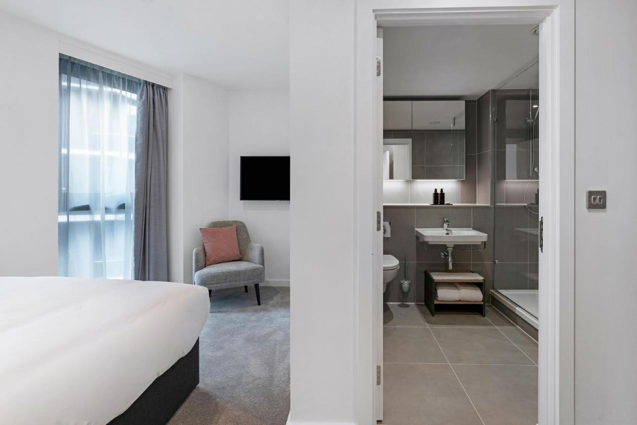 Citysuites 2 Aparthotel Manchester Exterior foto The photo shows a modern hotel room and bathroom setup. On the left, there is a bed with white bedding and a small sitting area that includes a gray chair and a pillow in a subtle pink shade. A flat-screen TV is mounted on the wall, providing enterta