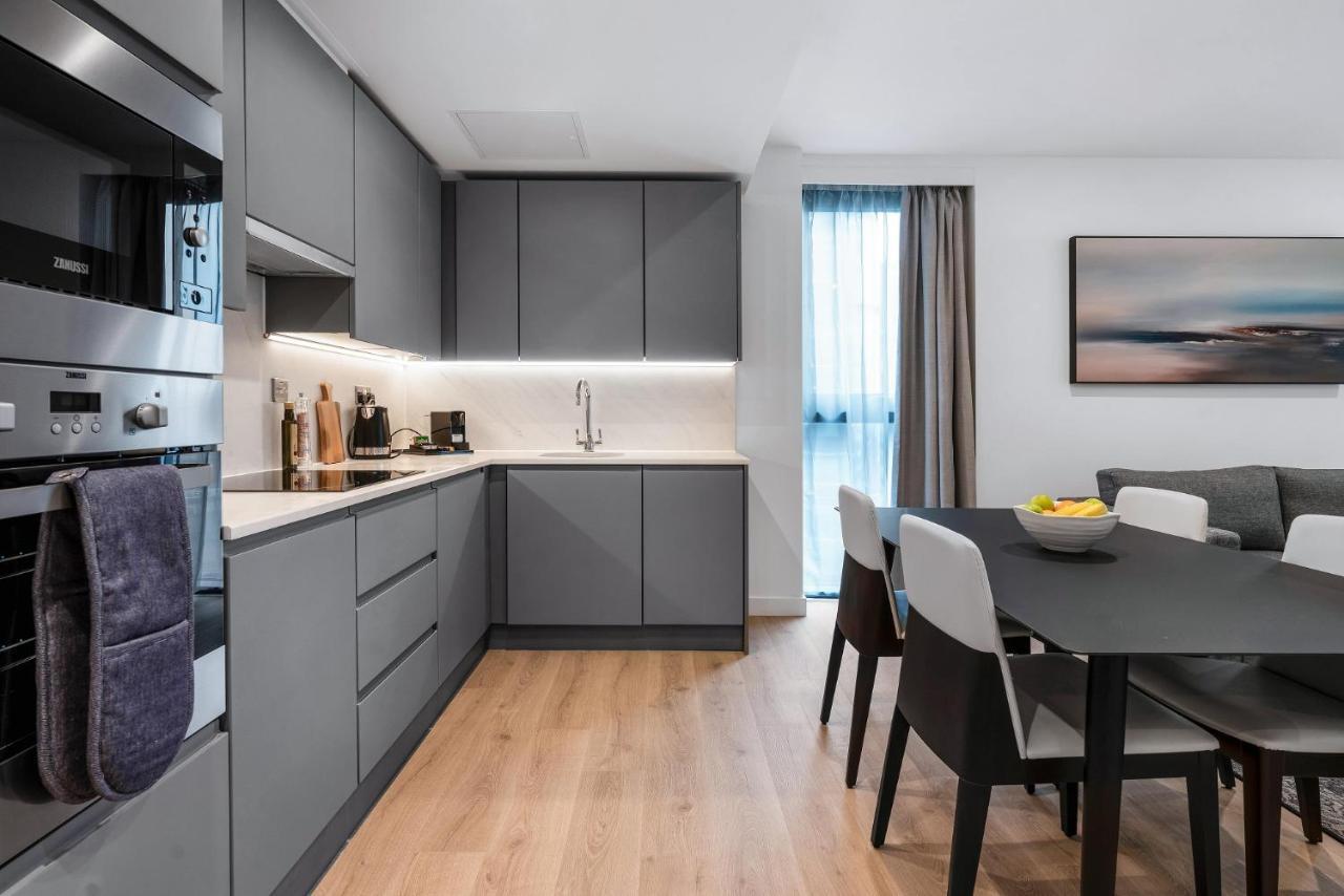 Citysuites 2 Aparthotel Manchester Exterior foto The photo shows a modern kitchen and dining area. The kitchen features sleek gray cabinetry, a white countertop, and built-in appliances such as an oven. There is a sink and some kitchen utensils displayed. The adjacent dining area contains a black t