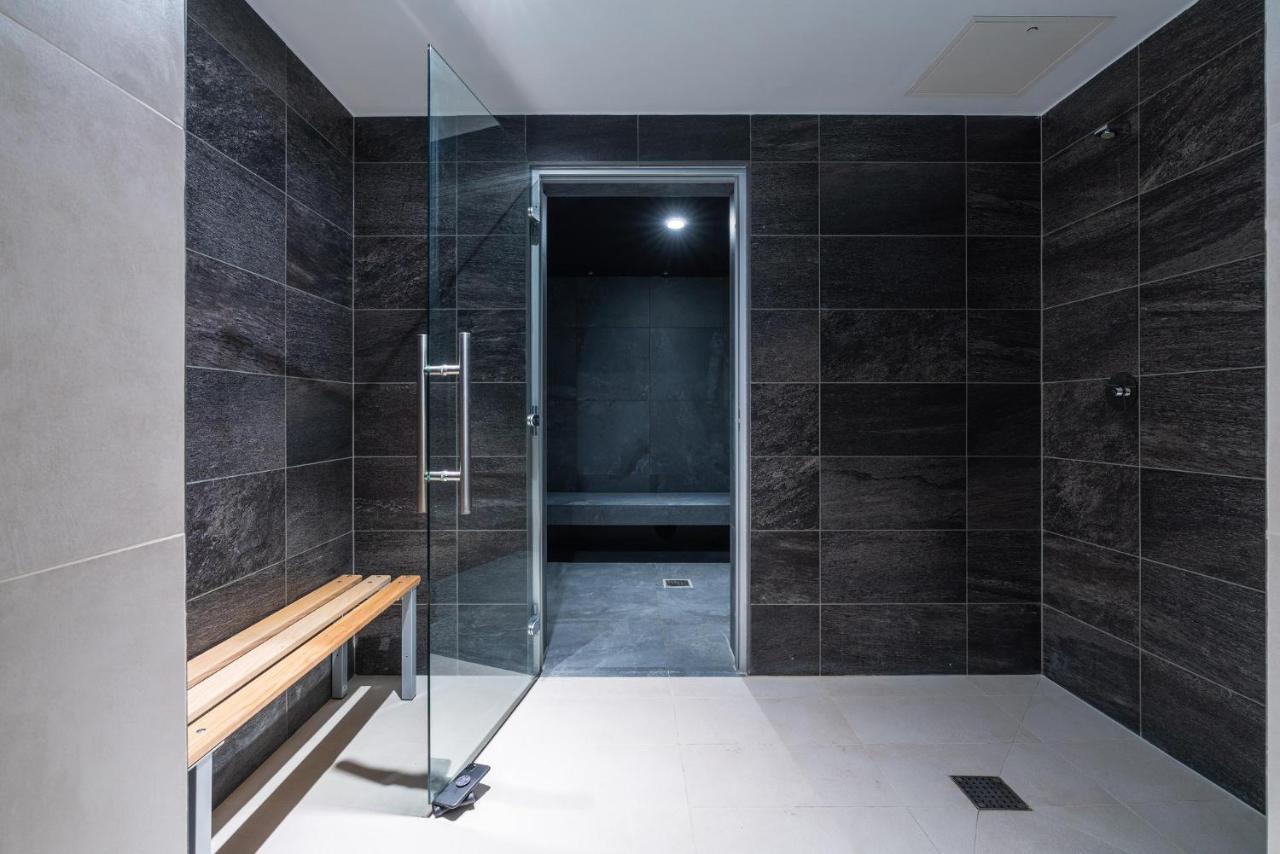 Citysuites 2 Aparthotel Manchester Exterior foto The photo shows a modern, minimalist shower area. The space is predominantly tiled with dark gray tiles on the walls, giving it a sleek and sophisticated look. There is a glass door that leads to another room, and a wooden bench is positioned against