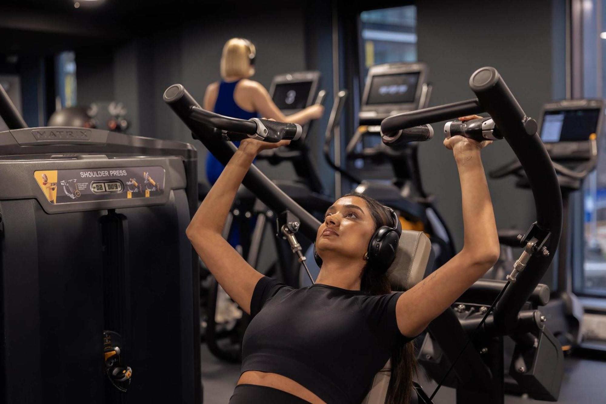 Citysuites 2 Aparthotel Manchester Exterior foto The photo shows a gym setting with two individuals engaged in physical exercise. In the foreground, a woman is using a chest press machine while wearing headphones, focusing on her workout. She appears to be in a seated position, pushing the handles 