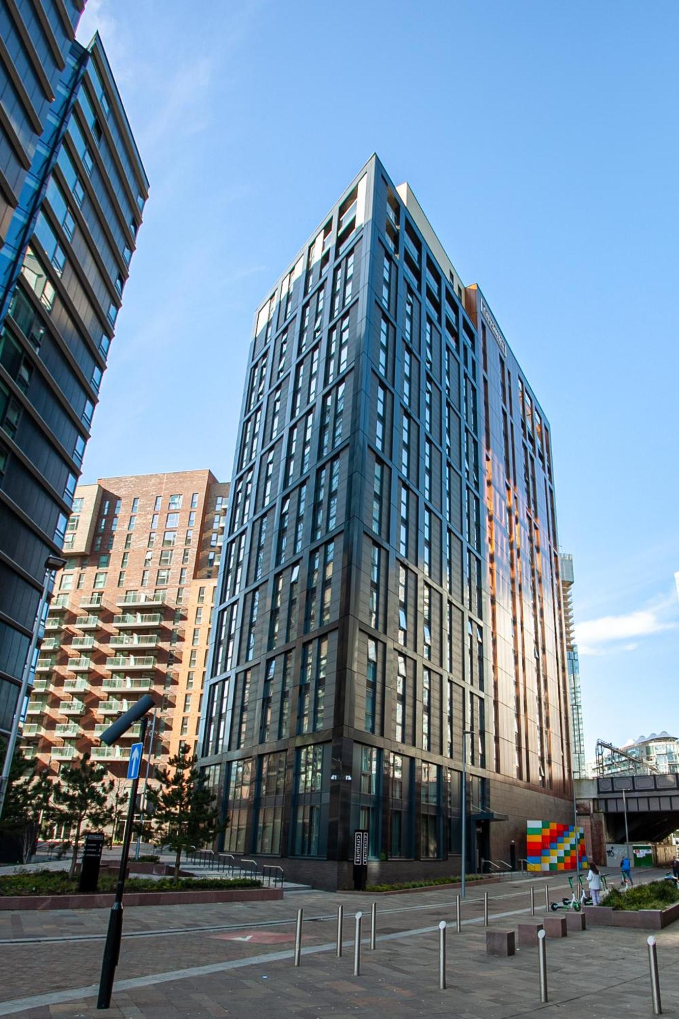 Citysuites 2 Aparthotel Manchester Exterior foto The photo shows a modern high-rise building with a sleek, glass façade. It is set against a blue sky, indicating a clear day. The building appears to have multiple floors with large windows that reflect light. Nearby, there are additional buildings, 